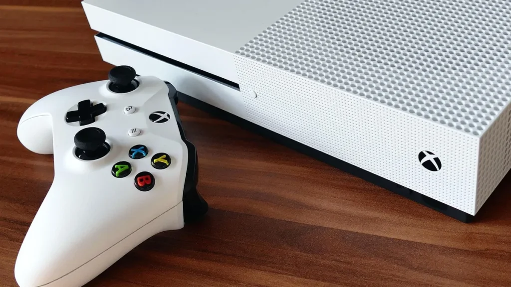 A white Xbox console and its controller lie on a polished wooden table.
