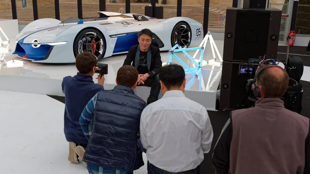 Kazunori Yamauchi is interviewed in front of a futuristic white and blue Alpine Vision GT concept car.